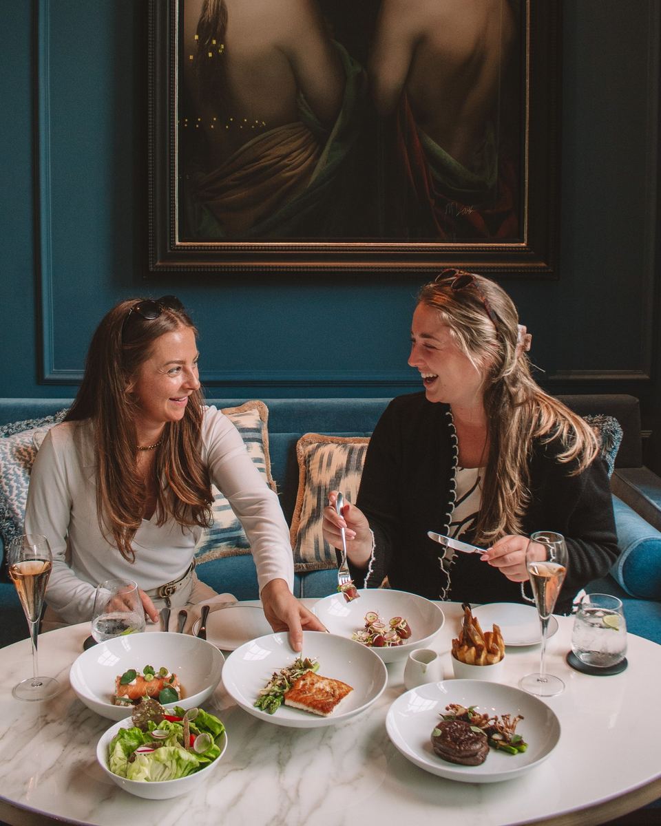 Two women laughing over dinner in a teal velvet booth